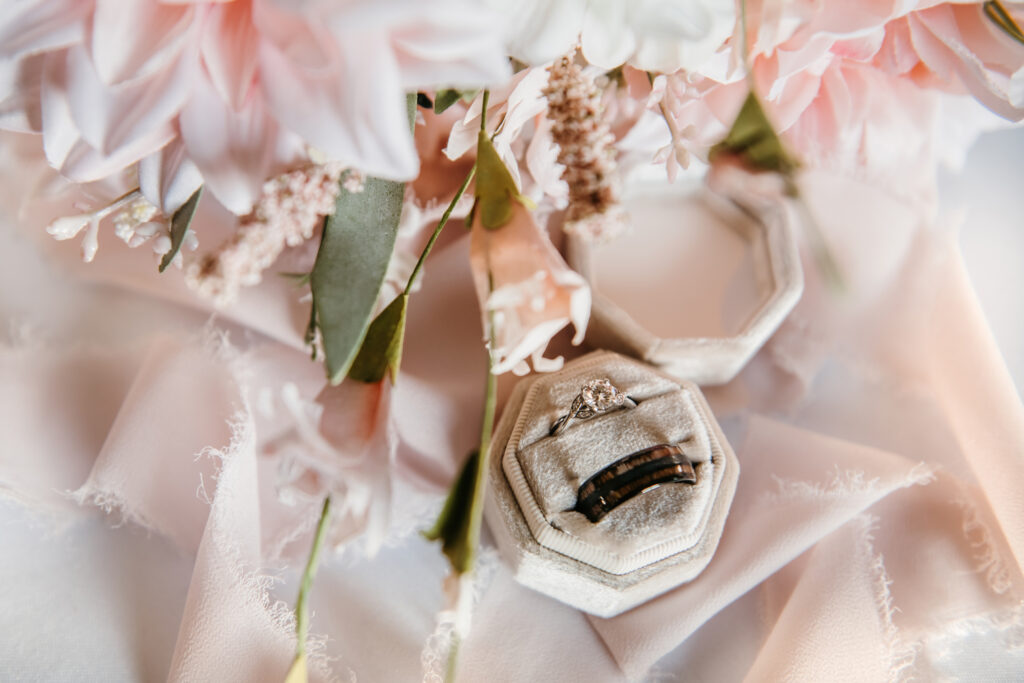 Pink flowers and ribbon surrounding a bride and groom's wedding rings in a cream ring box