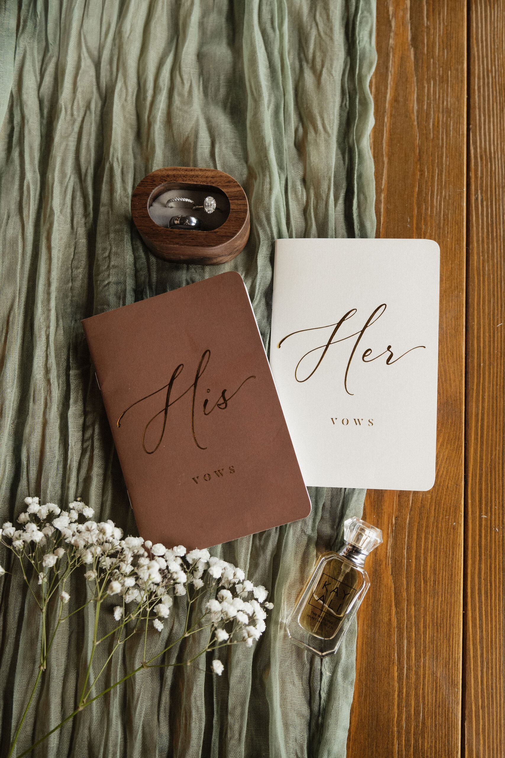 Two vow books placed next to each other with a wooden ring box, green cloth, and babies breath flowers around them.