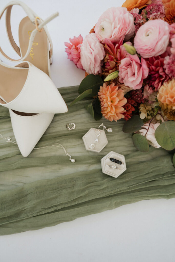 Green table runner with wedding jewelry, shoes, and pink and orange flowers laid out on it.