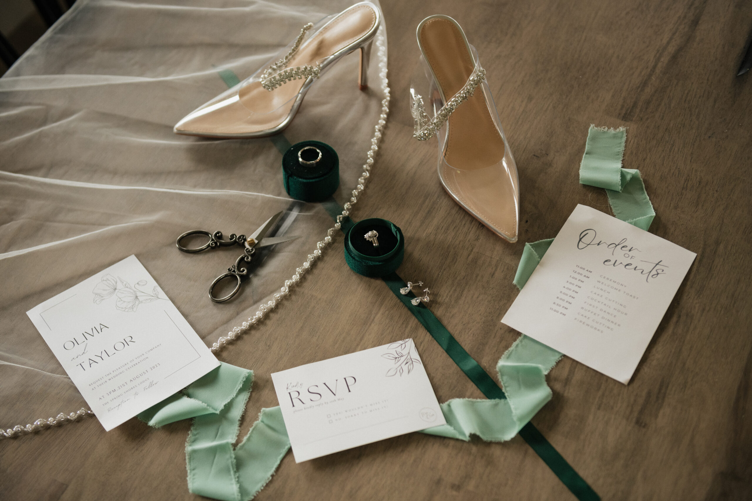 Green flatlay of wedding details, including a pair of heels, a ring box, a ribbon, earrings, and fabric scissors.