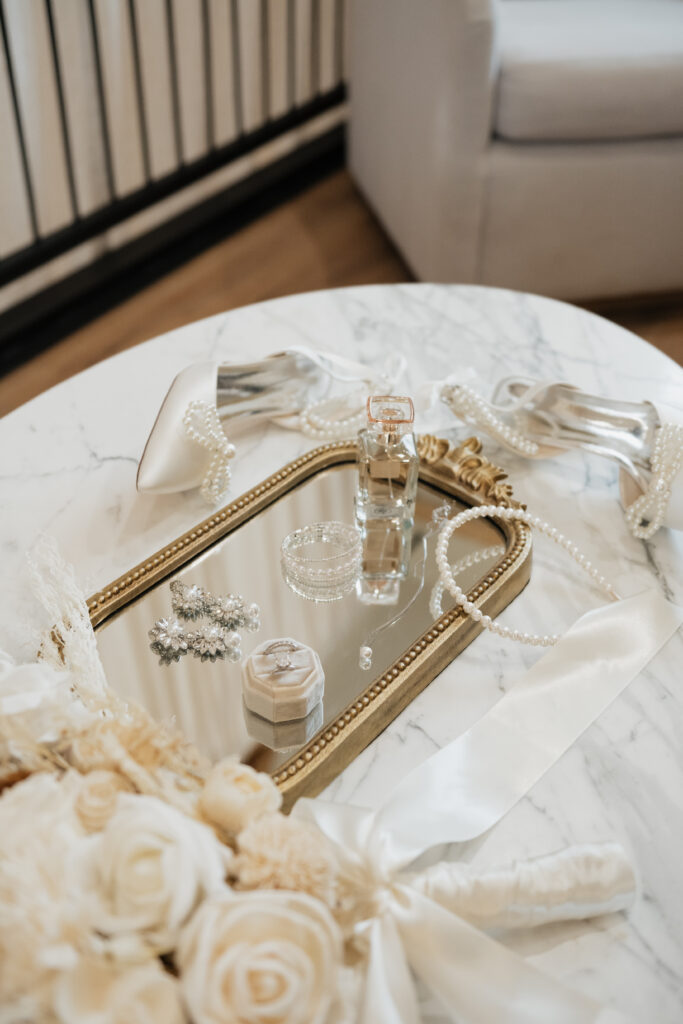 White and gold wedding flatlay setup with all the the bride's jewelry, perfume, pearls, and a pair of heels